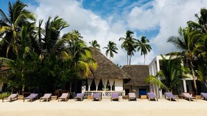 Um resort à beira-mar com edifícios de telhado de palha, palmeiras e espreguiçadeiras.