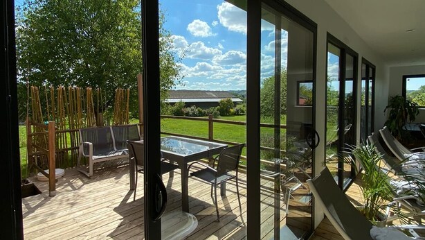 Eine moderne Außenterrasse mit Glastür zum Patio, inklusive Tisch und Stühlen, mit Blick auf eine grüne Landschaft.