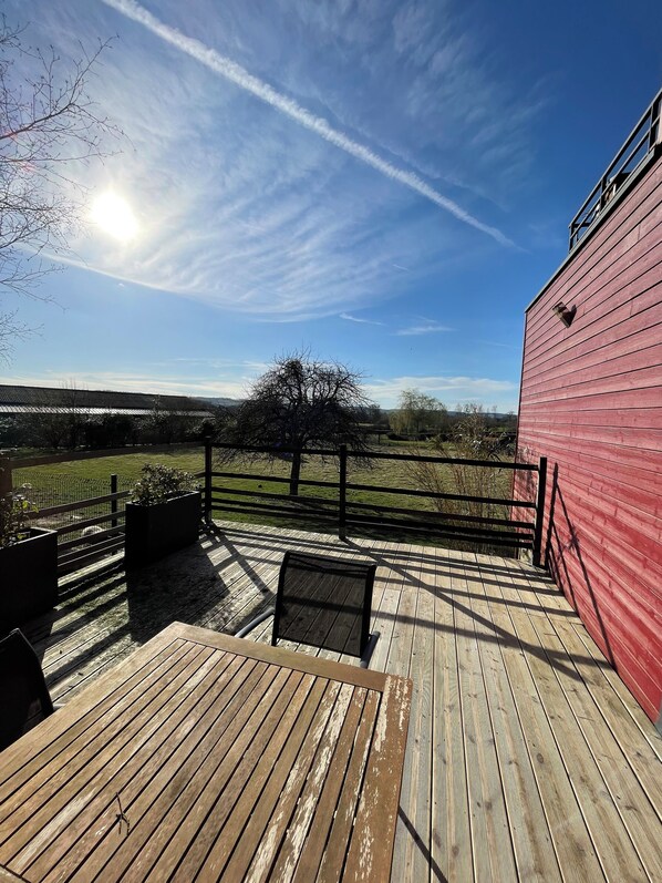Una terraza de madera con baranda metálica, un edificio rojo y vistas a un campo verde y un árbol.