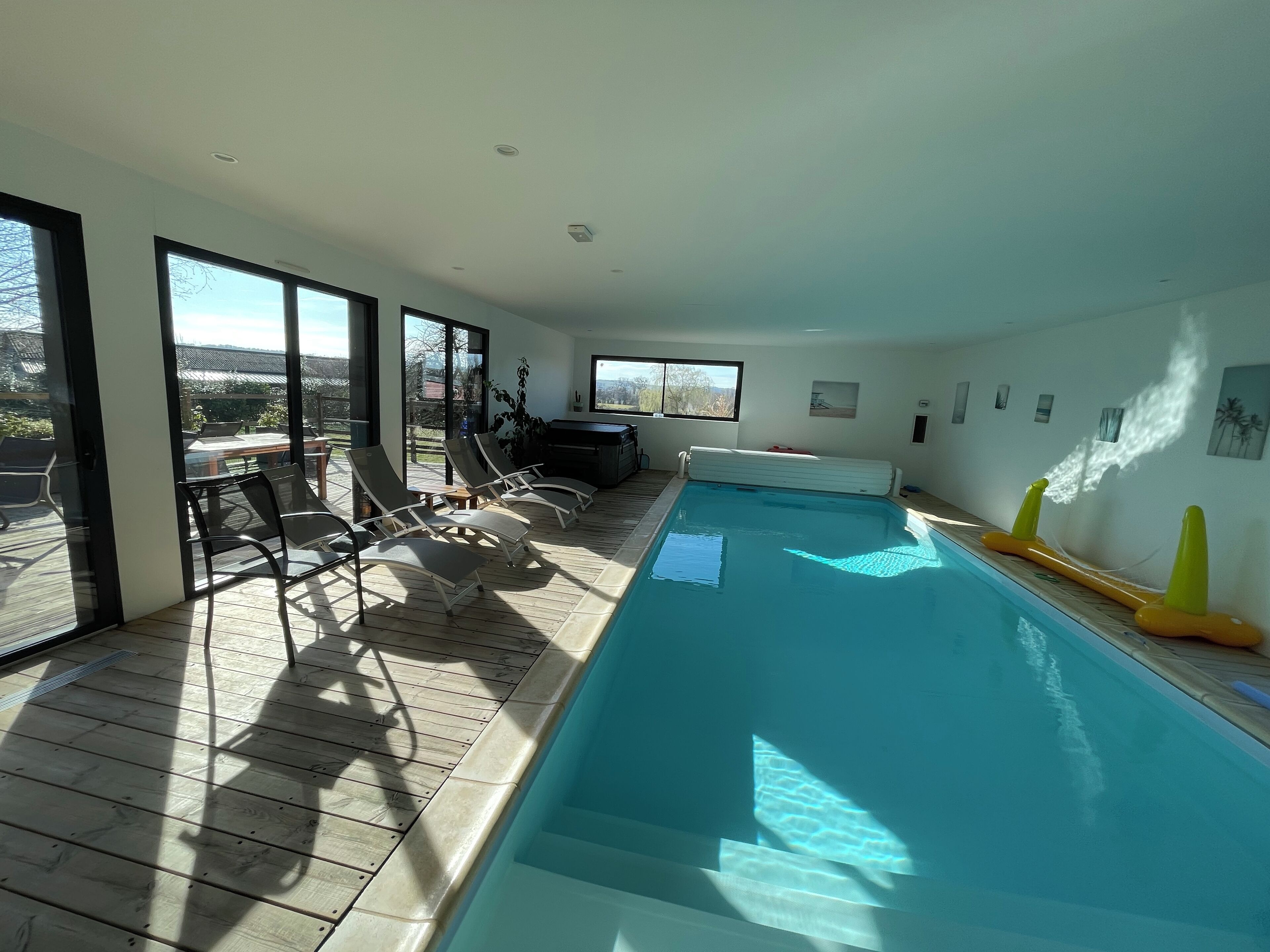 A modern indoor swimming pool with clear glass walls, lounge chairs, and a hot tub on an upper deck.