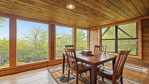 Una sala da pranzo in legno con un tavolo grande e sedie, ampie finestre che si affacciano su alberi e un soffitto in legno.