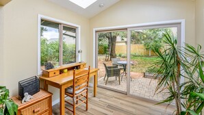 Ein Zimmer mit einem Holztisch, einem Stuhl und einer großen Glasschiebetür, die zu einem Garten mit Terrasse und Baum führt.