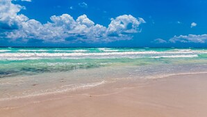 Une plage aux eaux turquoise et limpides, bordée d’un rivage sablonneux, sous un ciel d’un bleu éclatant parsemé de quelques nuages.