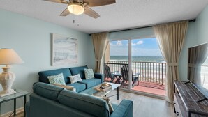 A living room with a sofa, coffee table, and a ceiling fan. There is a balcony with a view of the ocean.
