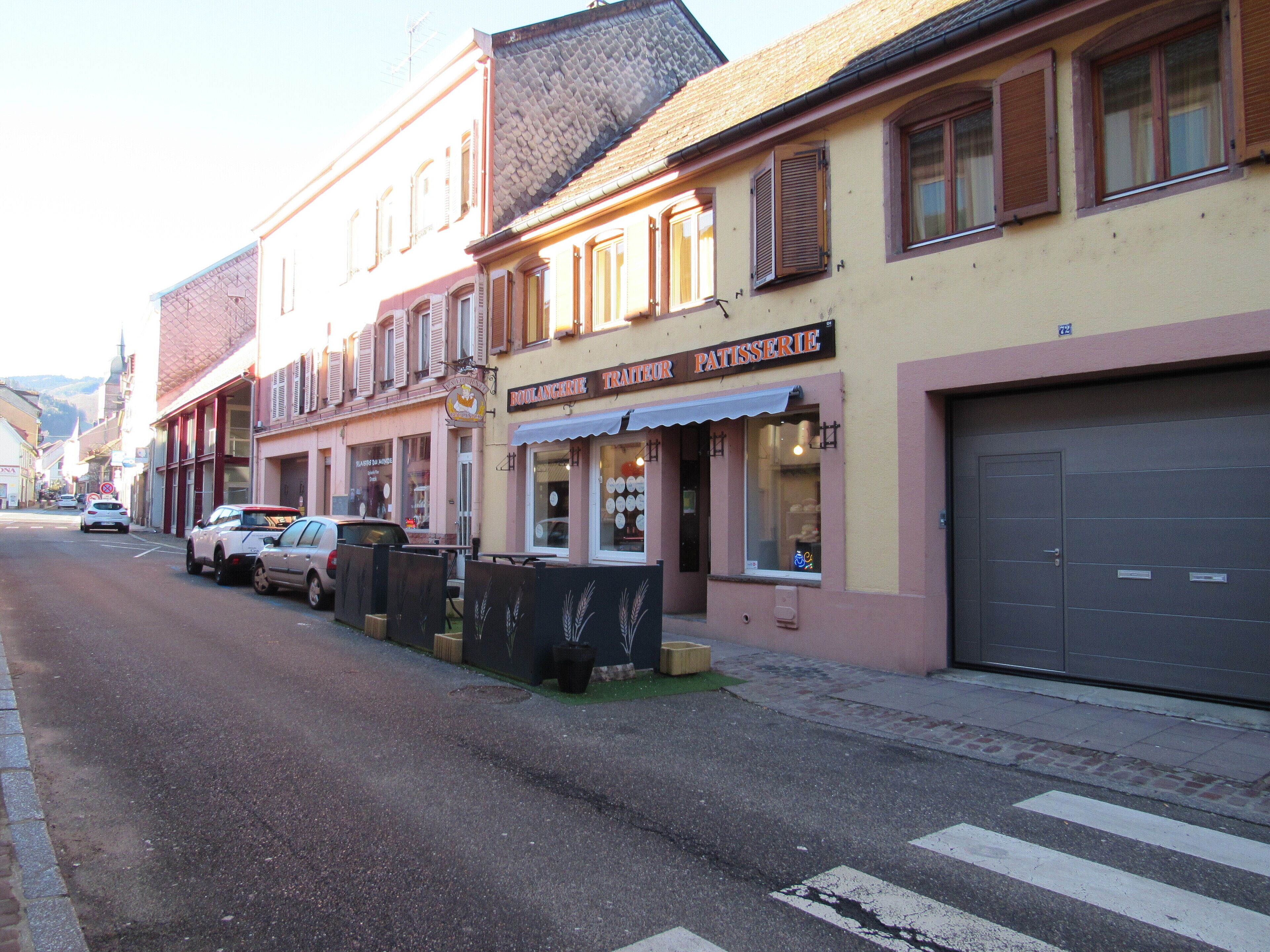 Une vue de rue avec une boulangerie-pâtisserie nommée « Boulangerie Pâtisserie » sur la droite, une voiture garée et un passage piéton.
