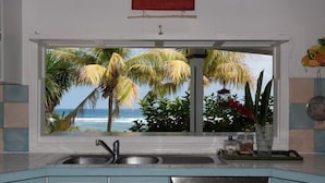 A kitchen with a large window overlooking a beach and palm trees.