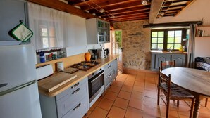 A kitchen with a stone wall, wooden ceiling beams, a dining table, and a window with a view of greenery. - Cottage on the Cevennes foothills with exceptional views (Sumene)