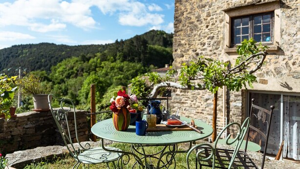 A rustic stone building with a small window, a round metal table with chairs, and a view of a green hillside. - Cottage on the Cevennes foothills with exceptional views (Sumene)