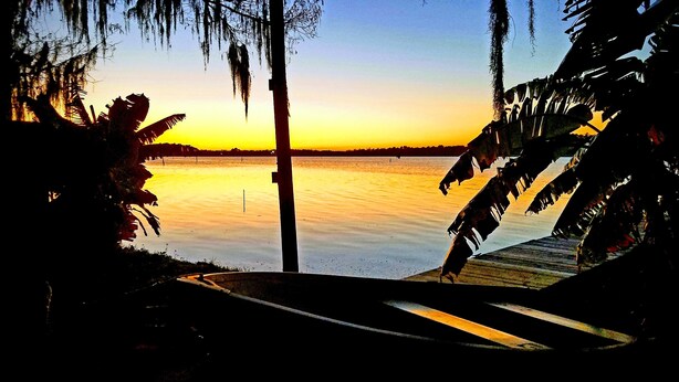 Une vue sur le coucher de soleil depuis un quai, avec une étendue d’eau calme et des palmiers.