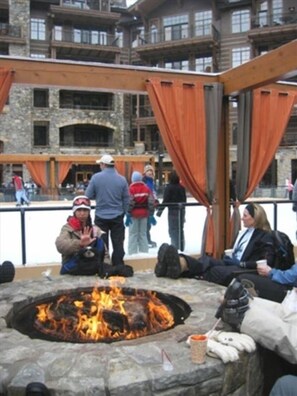 A stone fire pit with people around it, a wooden deck, and a building with stone walls and balconies in the background. - Northstar Ski Trail Condo Ski In/Ski Out NEWLY REMODELED (Truckee)