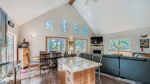 A spacious kitchen with a central island, wooden cabinets, and a dining area with chairs. There are large windows providing natural light and views of greenery.