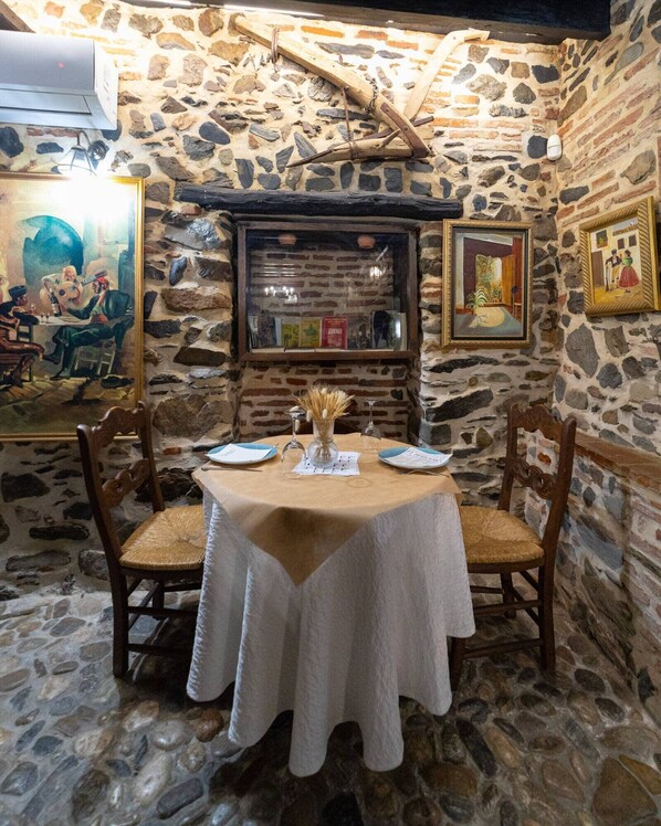 A stone-walled room with a table set for two, featuring a white tablecloth, two wooden chairs, and a vase with dried flowers. - Hotel Posada Del Bandolero (El Borge)