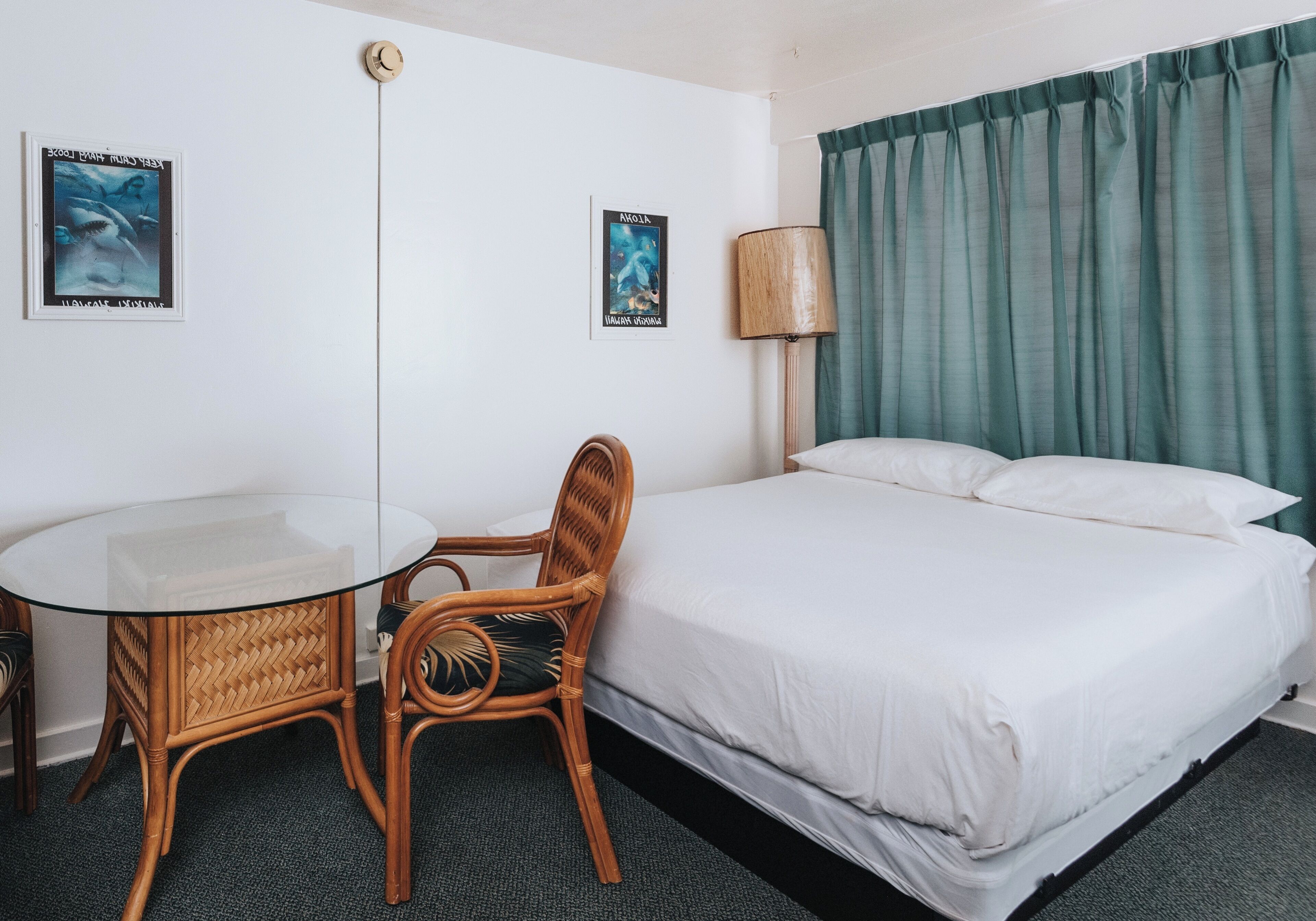 A hotel room with a bed, a round glass table, two wicker chairs, a lamp, and two posters on the wall.