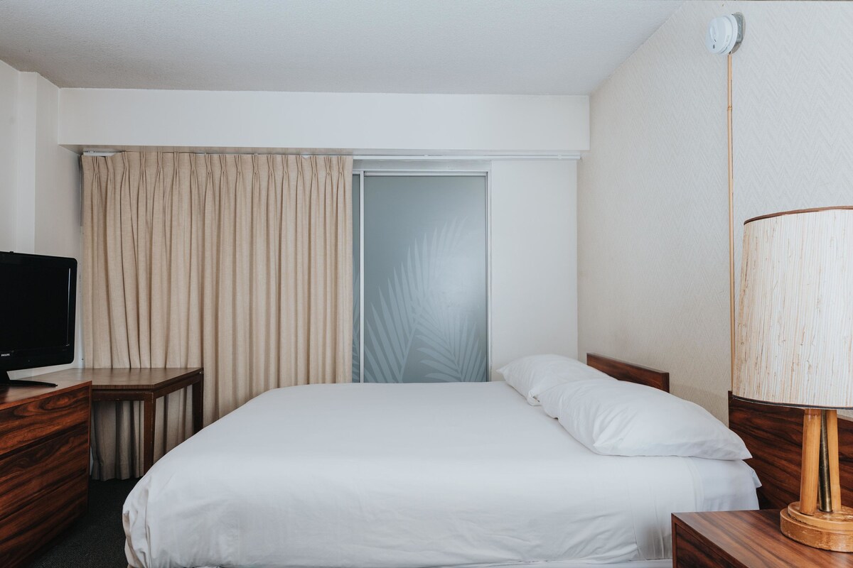 A hotel room with a bed, a television on a wooden nightstand, a lamp, and a sliding door.