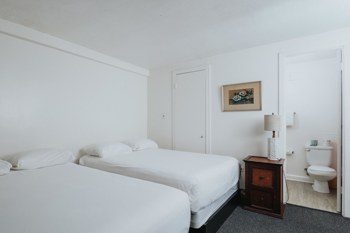 A hotel room with two single beds, a small wooden nightstand with a lamp, a framed picture on the wall, and a toilet visible through an open door.