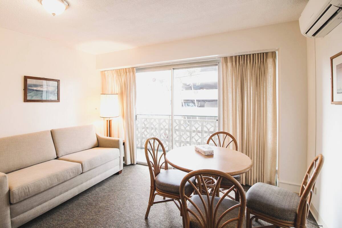 A room with a sofa, a round table with chairs, a lamp, and a window with curtains.