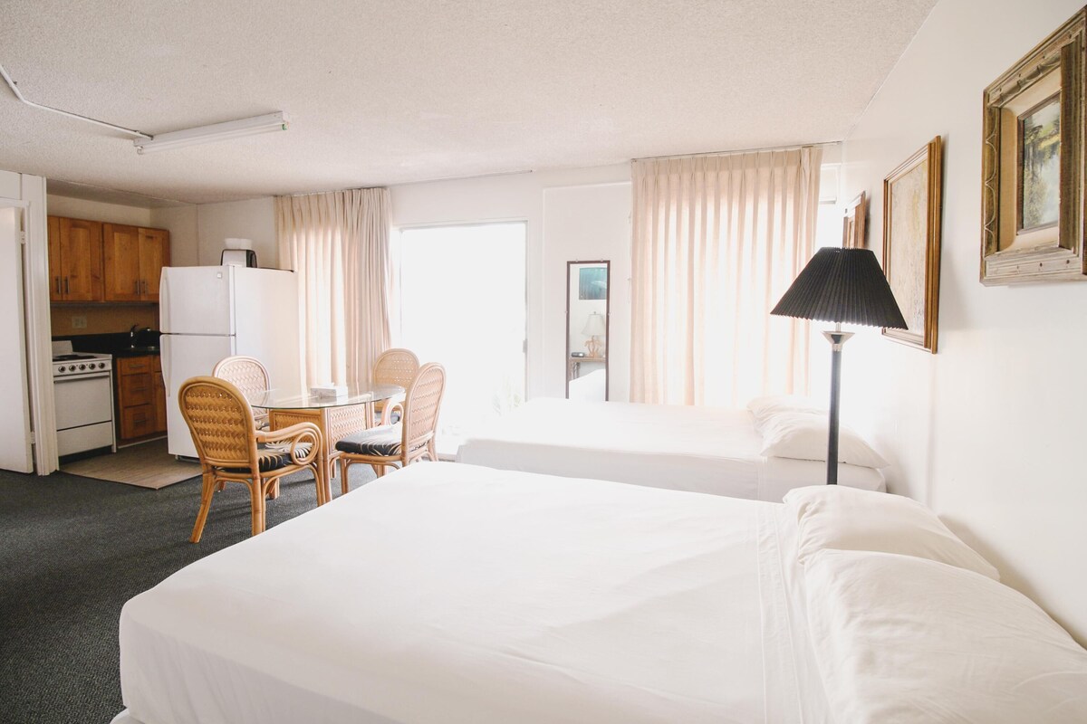 A hotel room with two beds, a dining table with chairs, a kitchenette, and a lamp.
