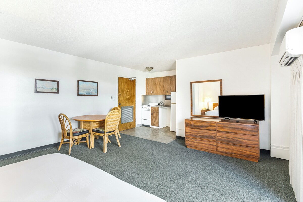 A hotel room with a bed, a TV, a dining table with chairs, a kitchen area, and artwork on the walls.