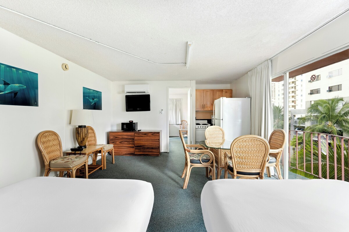 A hotel room with two beds, a dining table, chairs, a kitchen area, and a balcony with a view of palm trees.