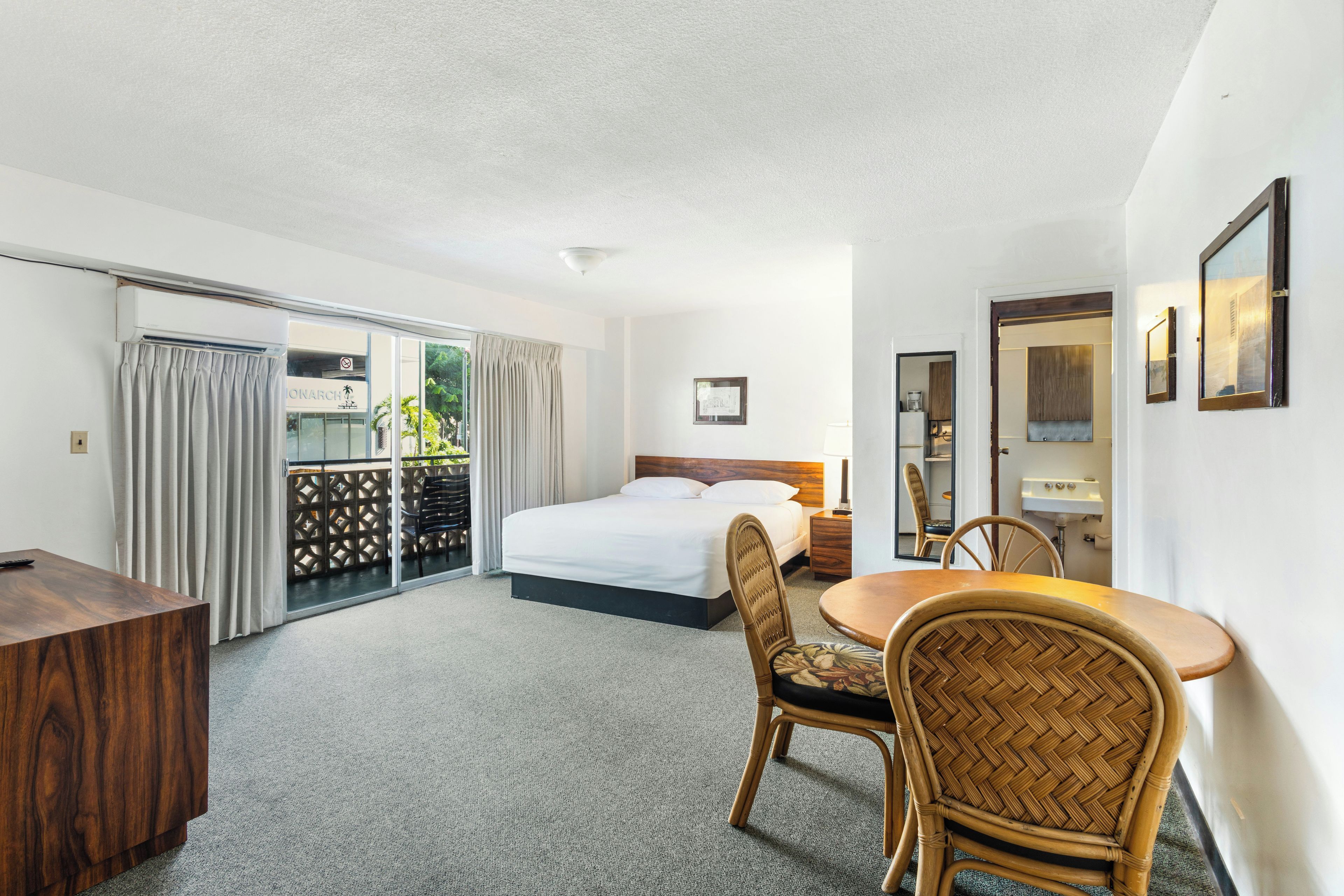 A hotel room with a bed, a dining table with chairs, a desk, a balcony with curtains, and a view of the outside.