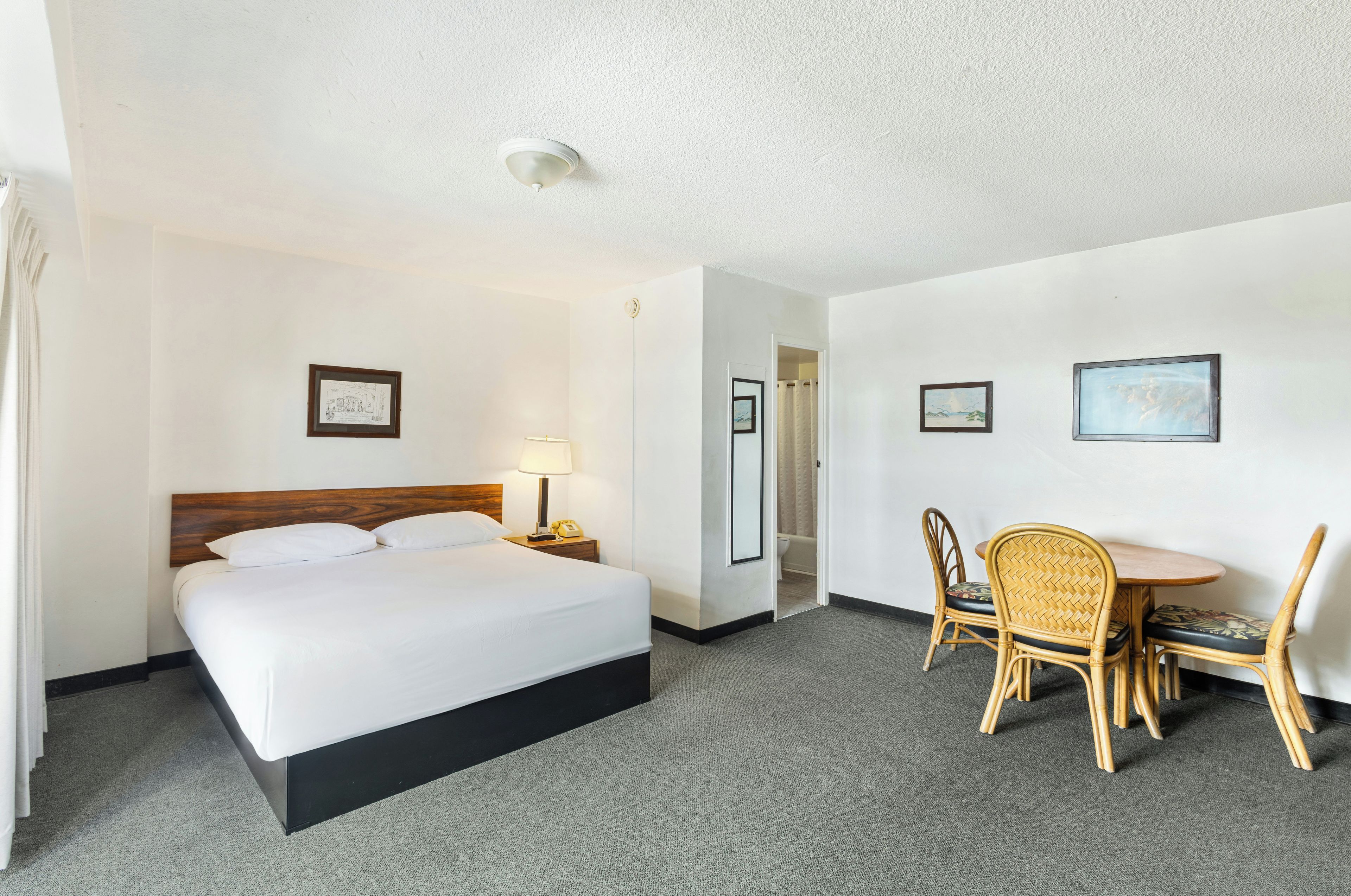 A hotel room with a bed, bedside lamp, a desk with chairs, and framed pictures on the wall.