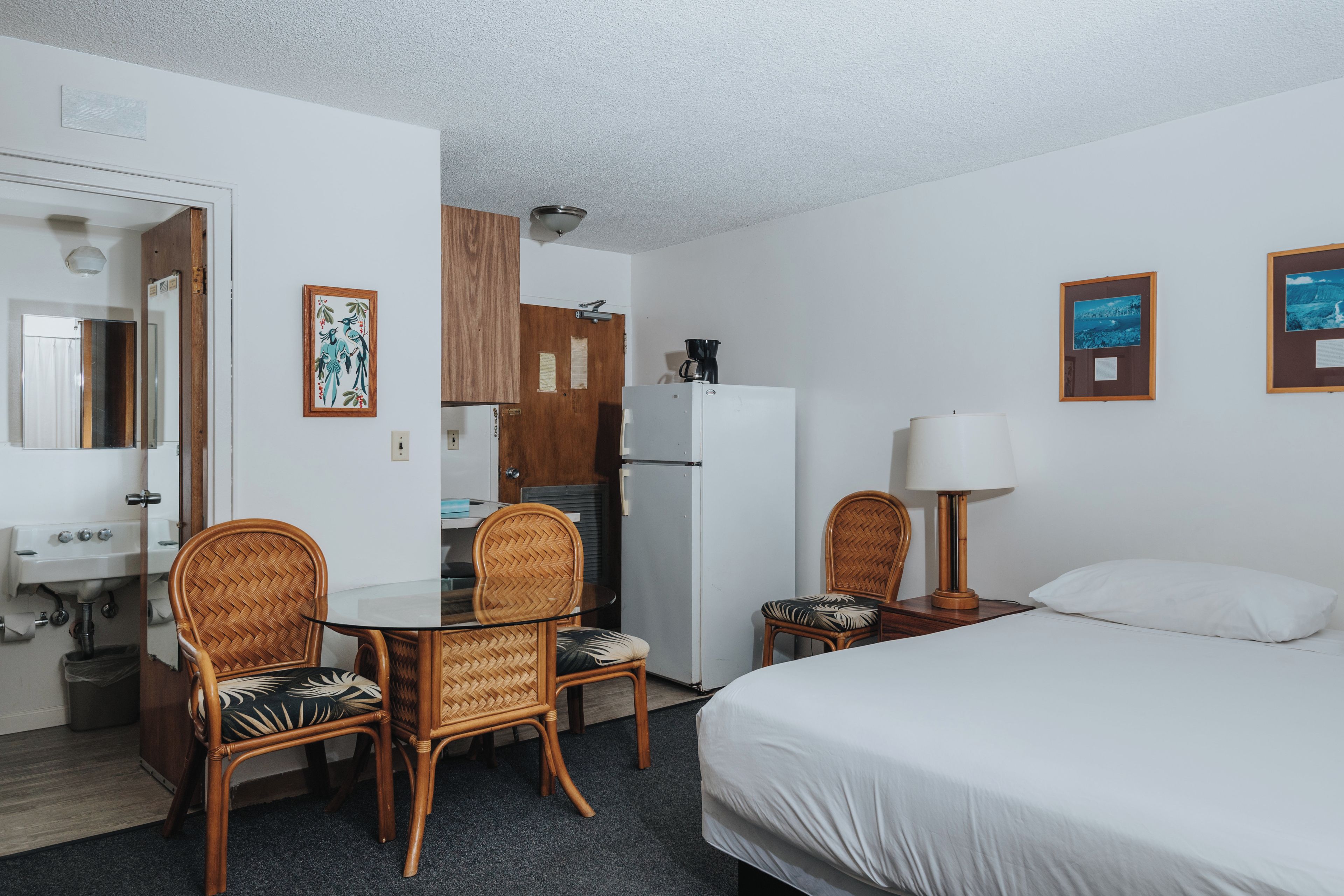 Une chambre d’hôtel comprenant un lit, deux fauteuils en osier, une petite table, un réfrigérateur, un lavabo et un miroir.