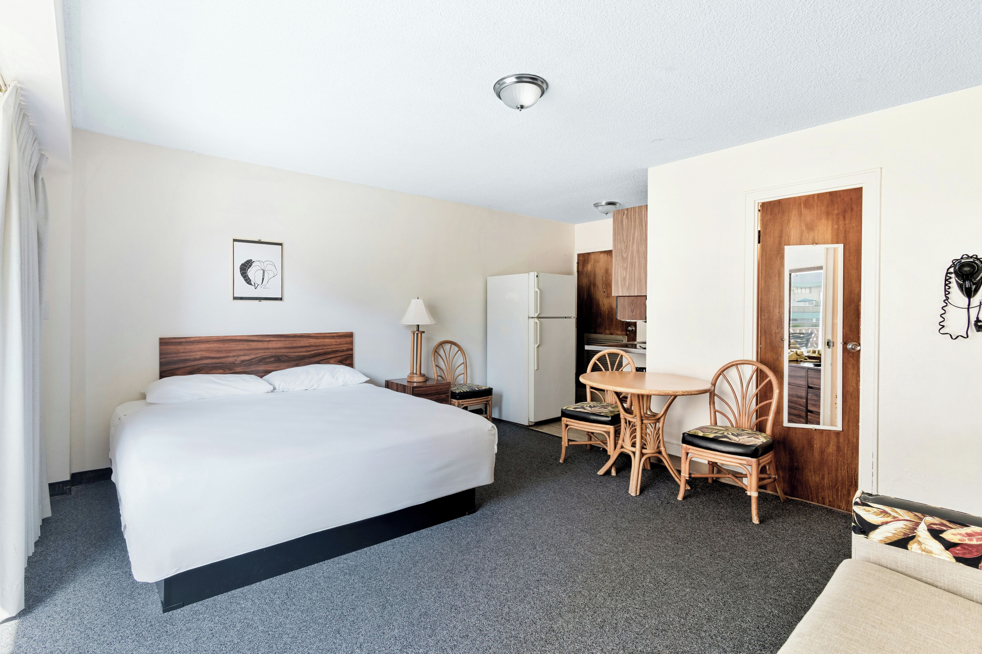 A hotel room with a bed, a desk, a chair, a lamp, a refrigerator, and a wooden door.