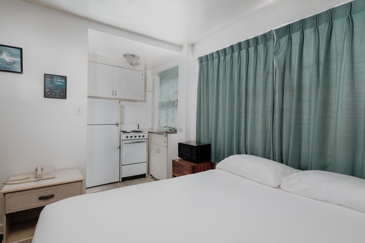 A compact hotel room with a bed, a kitchenette, a small table with a phone, and a television.