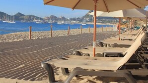 Una playa con sillas de descanso y sombrillas, arena dorada y veleros al fondo.