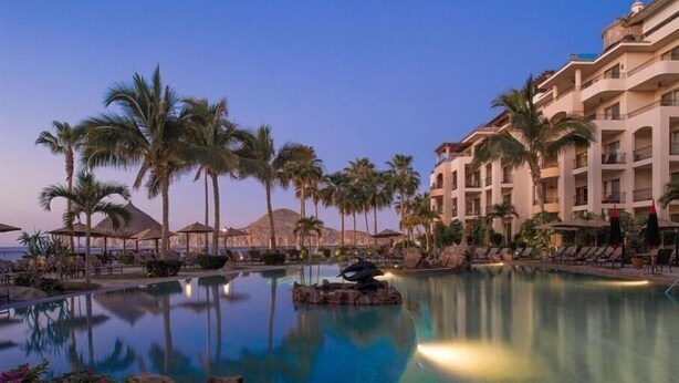 Piscina de resort con palmeras, sillas de descanso y un edificio con balcones.