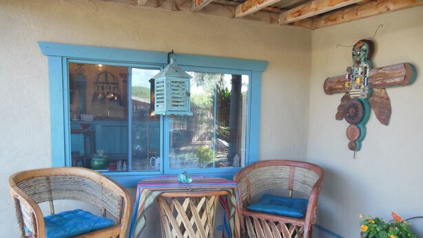 A small patio area with wicker furniture, a table with a colorful striped tablecloth, and a window with a view of greenery outside. -  Casita Juliet in Taos, New Mexico (Taos)