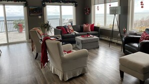 Un salon spacieux avec une grande fenêtre donnant sur la mer, un canapé gris agrémenté de coussins rouges, un fauteuil à carreaux et un petit pouf.