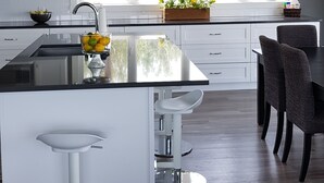 Une cuisine moderne avec des meubles blancs, un grand îlot central doté de tabourets, une fenêtre donnant sur la verdure, et des panneaux indiquant « BON APPÉTIT ! » et « LA CUISINE ».