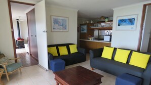 Een woonkamer met twee banken, een houten salontafel en een keuken met een koelkast en planken.