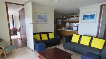 A living room with two sofas, a wooden coffee table, and a kitchen area with a refrigerator and shelves.