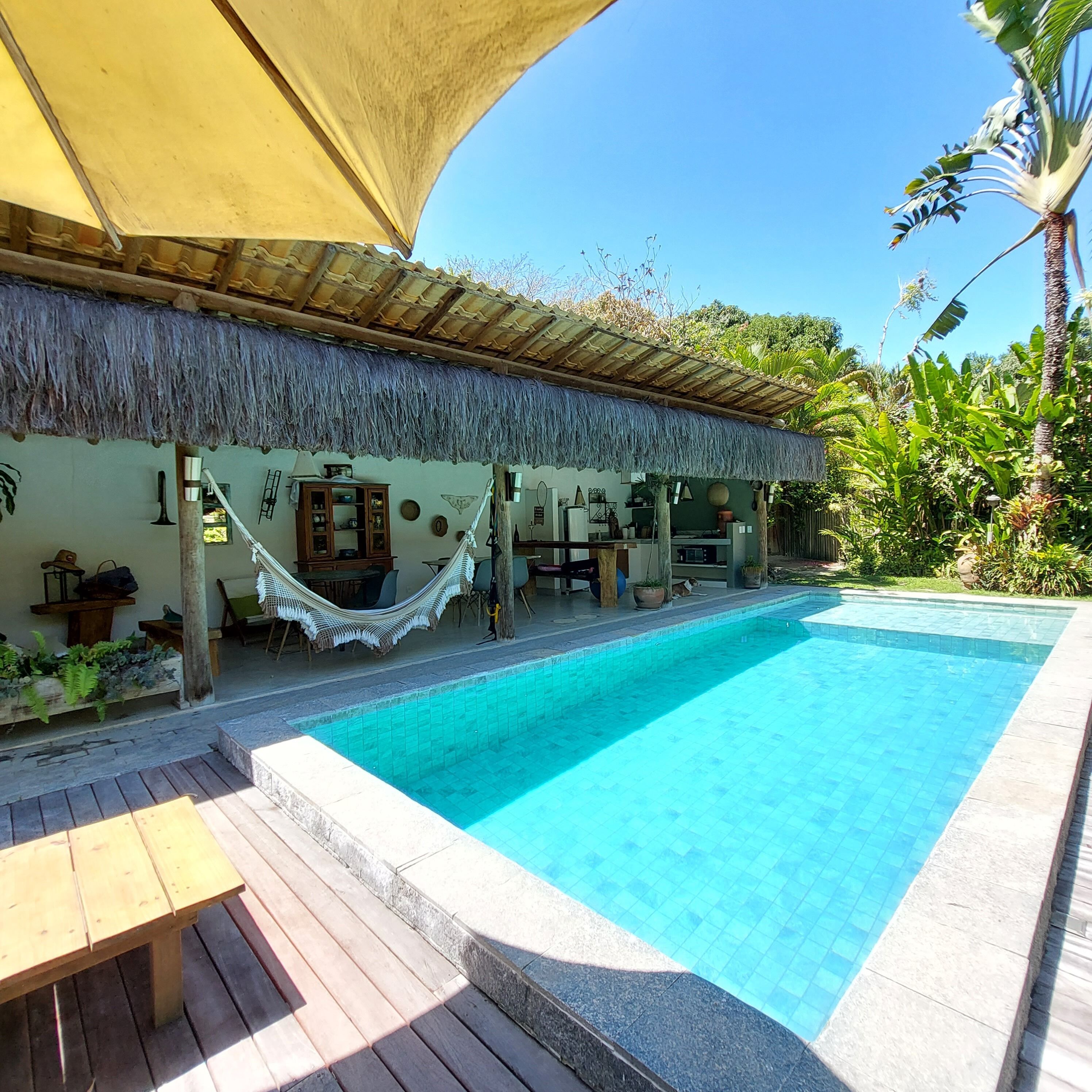 A pool area with a thatched roof structure, wooden deck, and a hammock.