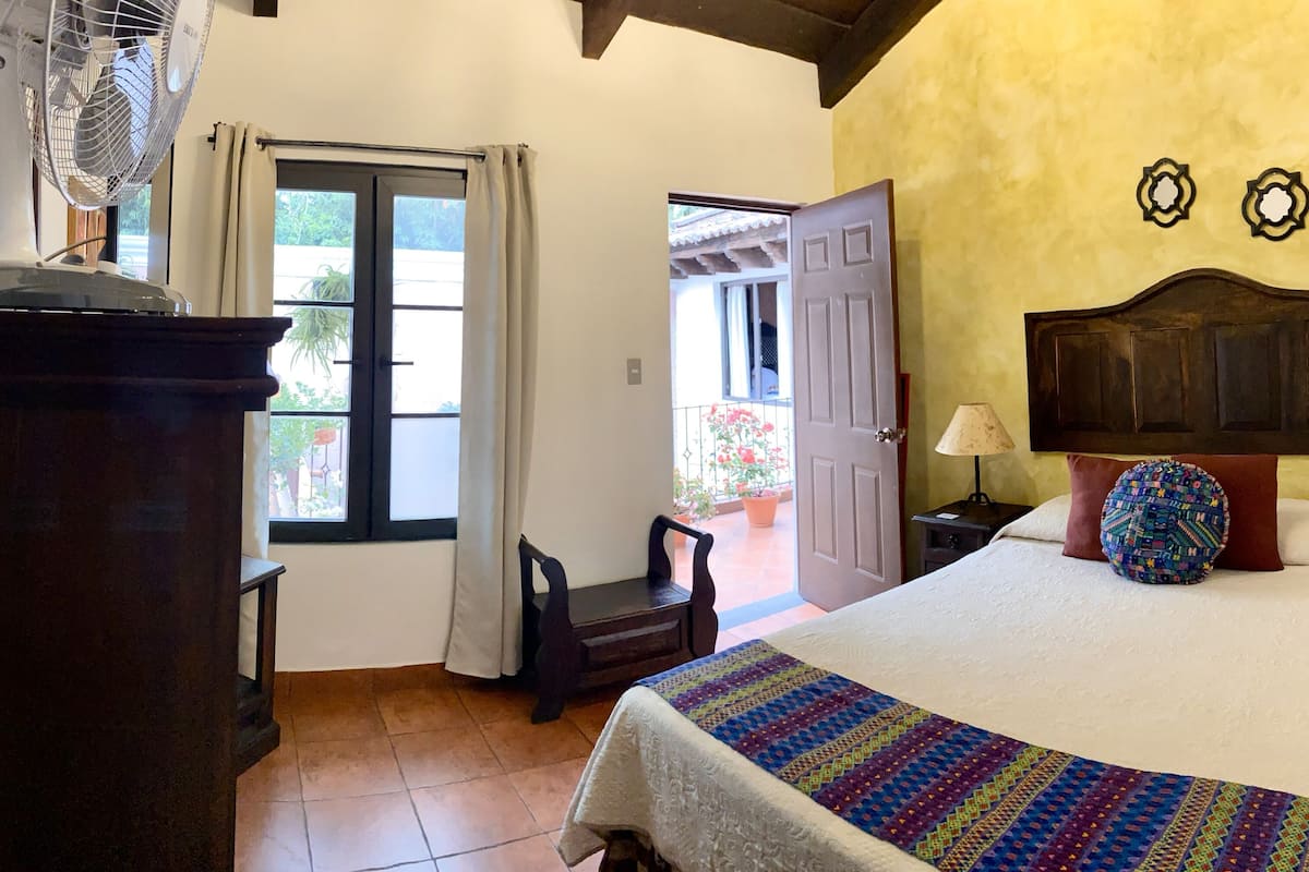 A bedroom with a bed, a nightstand, a lamp, a fan, a window with curtains, and a door leading to a balcony.