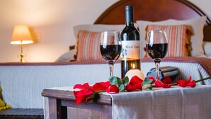 Una cama con cabecera de madera, una mesita de noche con una botella de vino y dos copas, y una vela encendida.