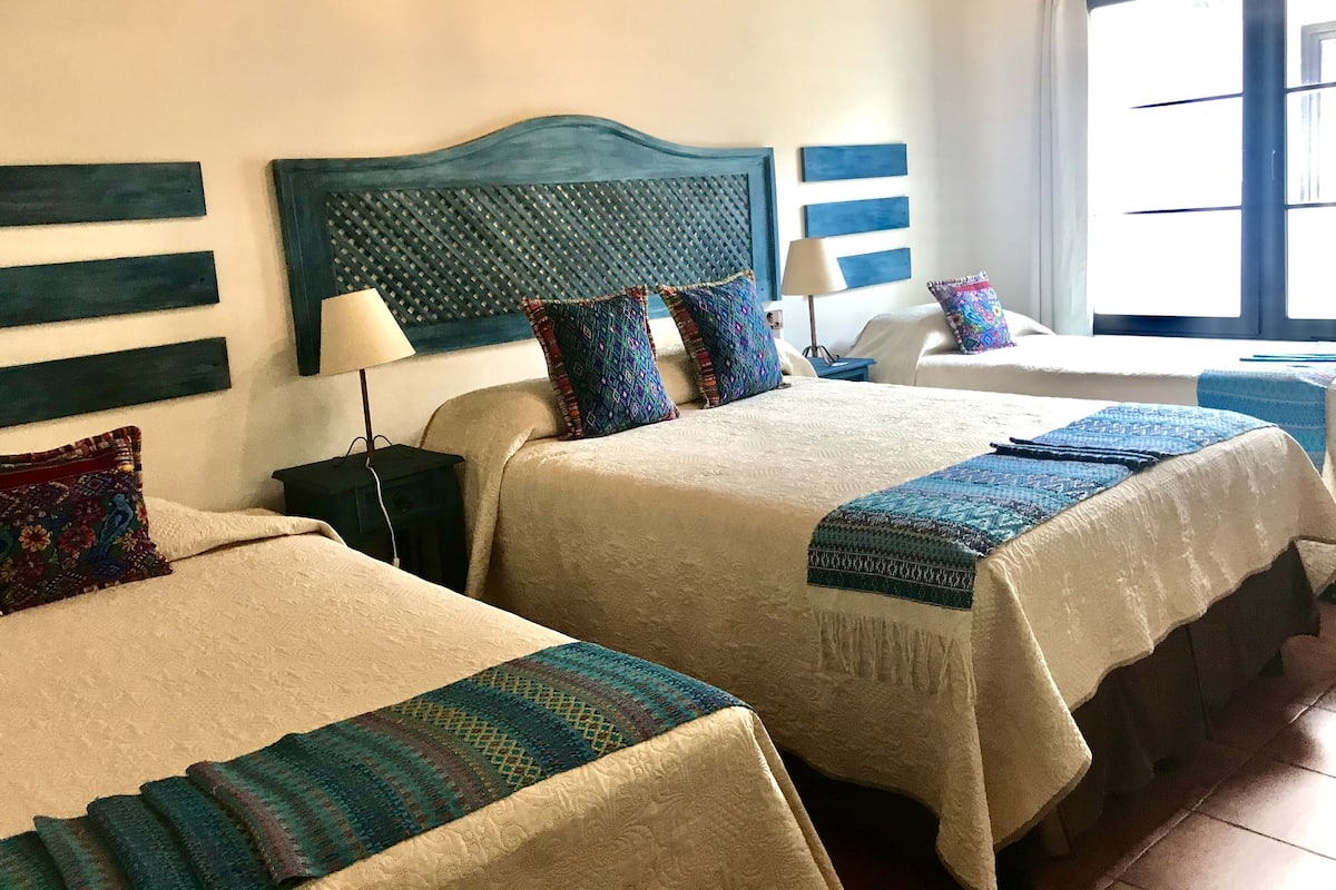 A hotel room with two beds, a headboard with a textured pattern, a nightstand with a lamp, and a window with blinds.