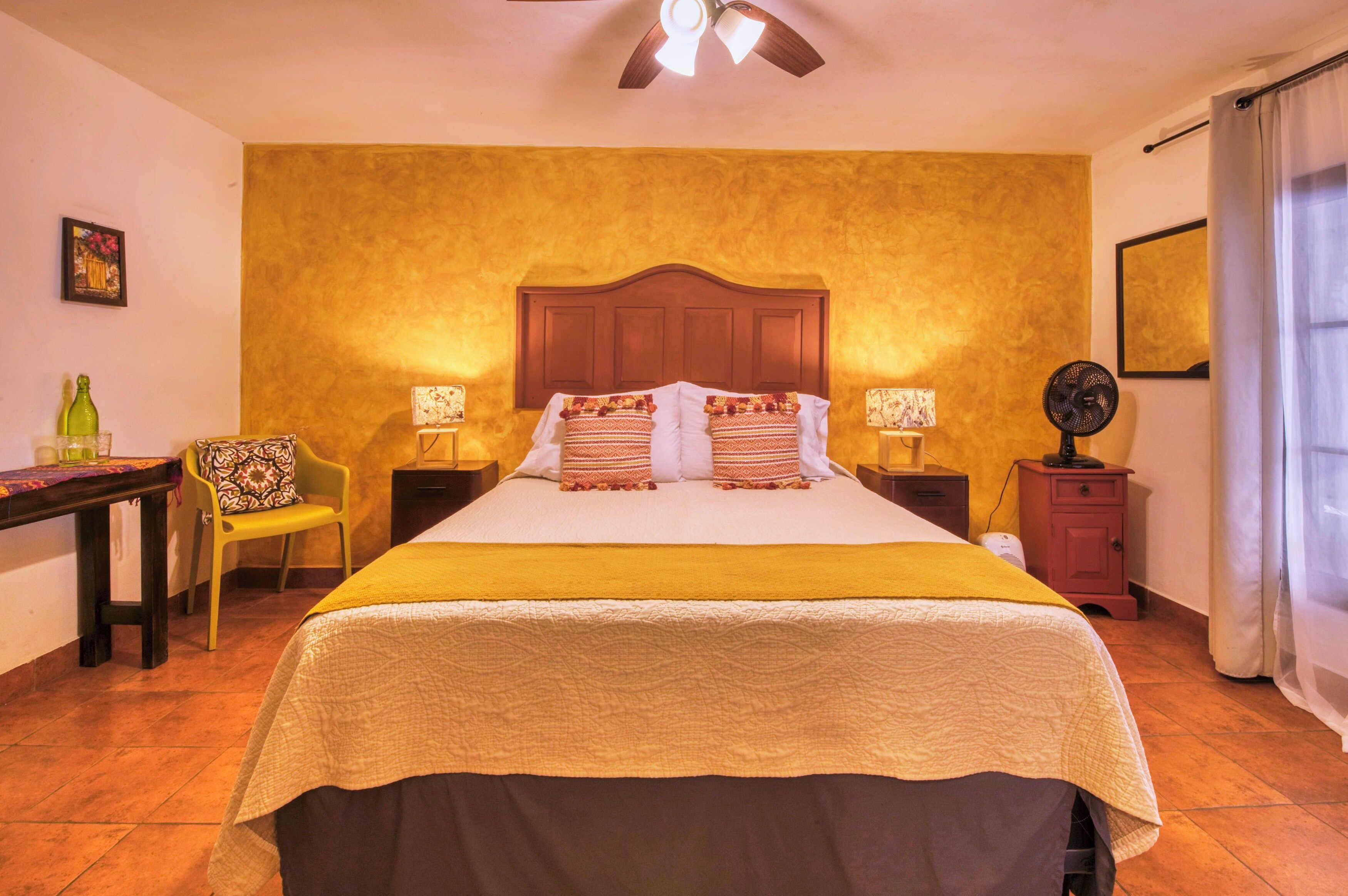 A hotel room with a bed, a nightstand, a chair, a desk, a lamp, a fan, and a ceiling fan.