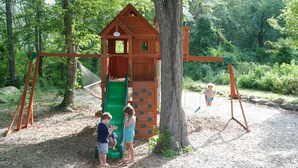 Une structure de jeux en bois, avec un toboggan et des balançoires, située dans un espace boisé.