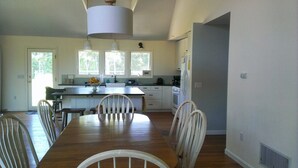 Une cuisine spacieuse dotée d’un îlot central, d’une table à manger en bois et de chaises. Trois fenêtres laissent entrer la lumière naturelle, et un grand luminaire suspendu est placé au-dessus de l’îlot.