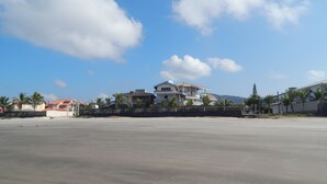 Uma casa grande, de vários andares, com telhado plano e varanda, cercada por uma vedação e palmeiras.
