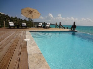 Eine Holzterrasse mit Pool, weißen Liegestühlen und Sonnenschirm.