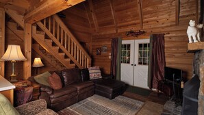 A log cabin interior with a wooden staircase, a leather sofa, a fireplace, and a patterned rug.