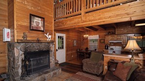 A log cabin interior with a stone fireplace, wooden walls, a dining area, and a cozy living room with a leather couch.