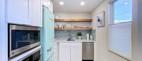 A compact kitchen with white cabinets, a blue refrigerator, stainless steel appliances, a tiled backsplash, and a window with blinds.