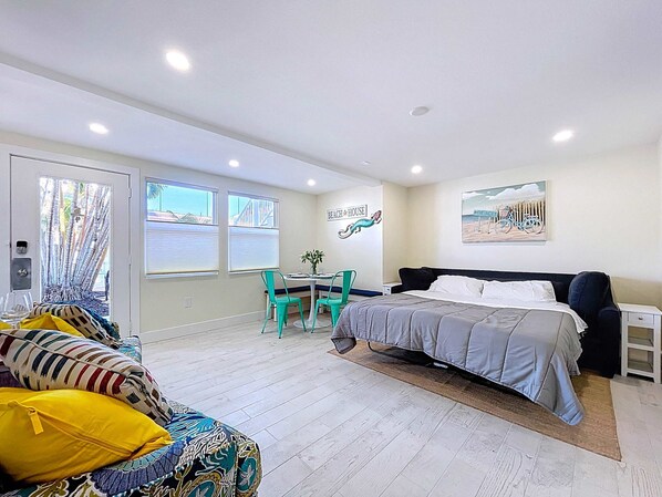 A bedroom with a bed, a desk, and a chair. There is a window with blinds, a door, and a wall with a painting.