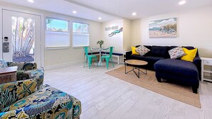A living room with a sofa, a dining table, and chairs. There is a painting on the wall and a window with blinds. - Bright Coastal Unit Steps From Beach/Walk 2 Bridge (bradenton beach)