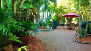 A paved pathway leading to a seating area with a red umbrella, surrounded by lush greenery and potted plants. - Bright Coastal Unit Steps From Beach/Walk 2 Bridge (bradenton beach)